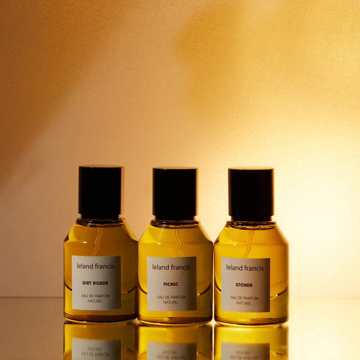 Three Leland Francis eau de parfum bottles labeled Dirt Roads, Picnic, and Stoner in warm light