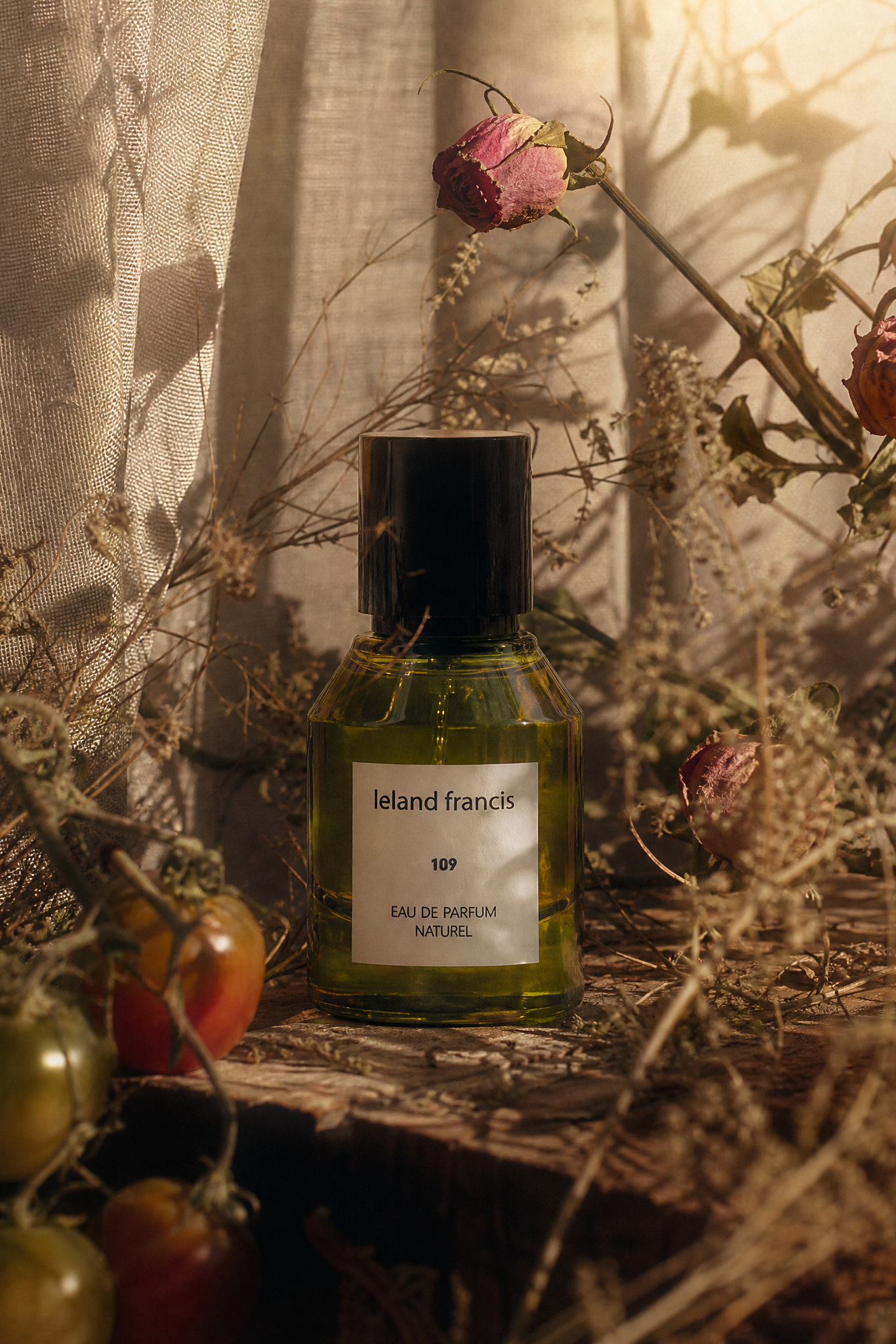 Perfume bottle labeled 'Leland Francis' surrounded by dried flowers and tomatoes on a textured surface.