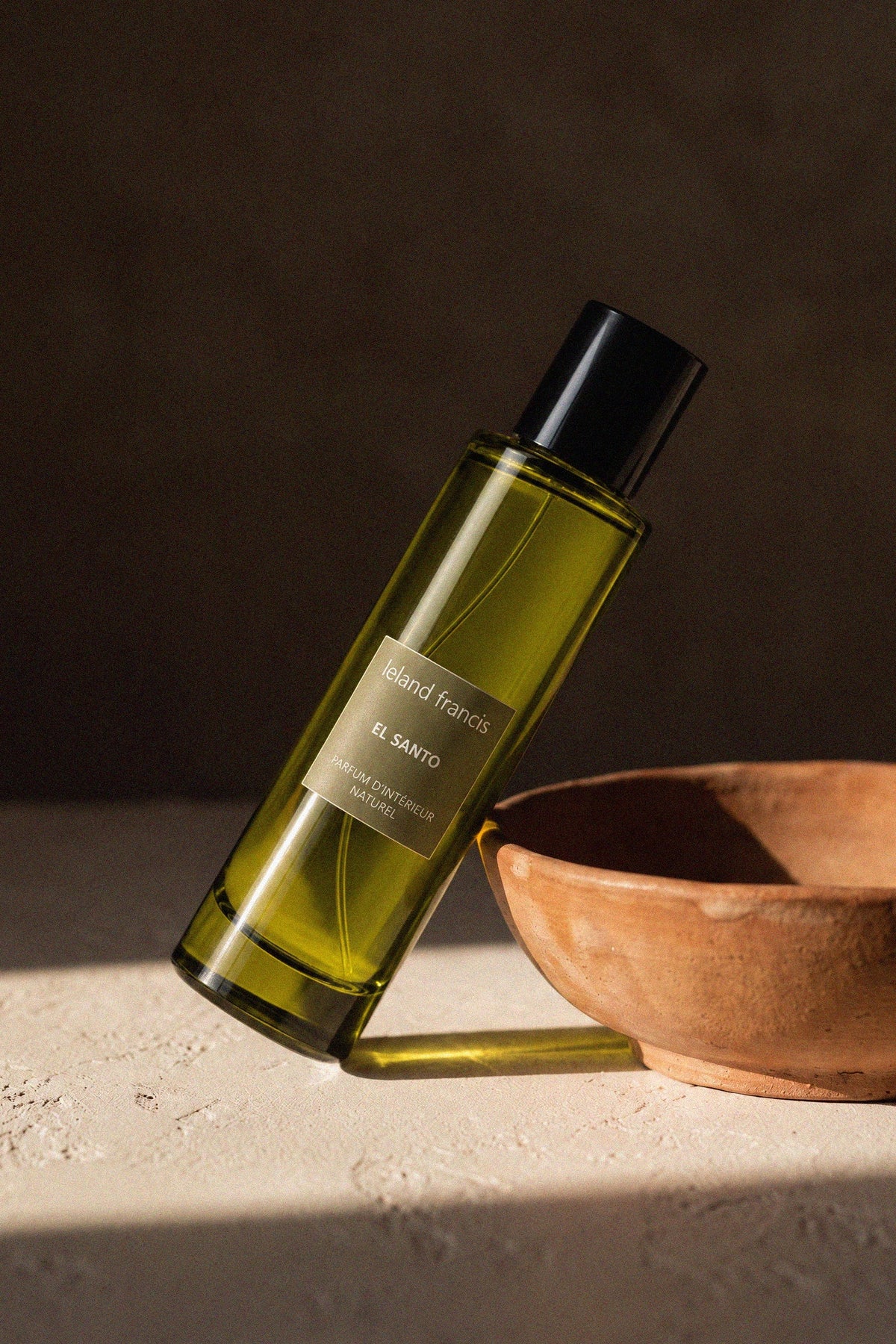 El Santo Room Spray leaning against a terracotta bowl on textured plaster, raking sunlight