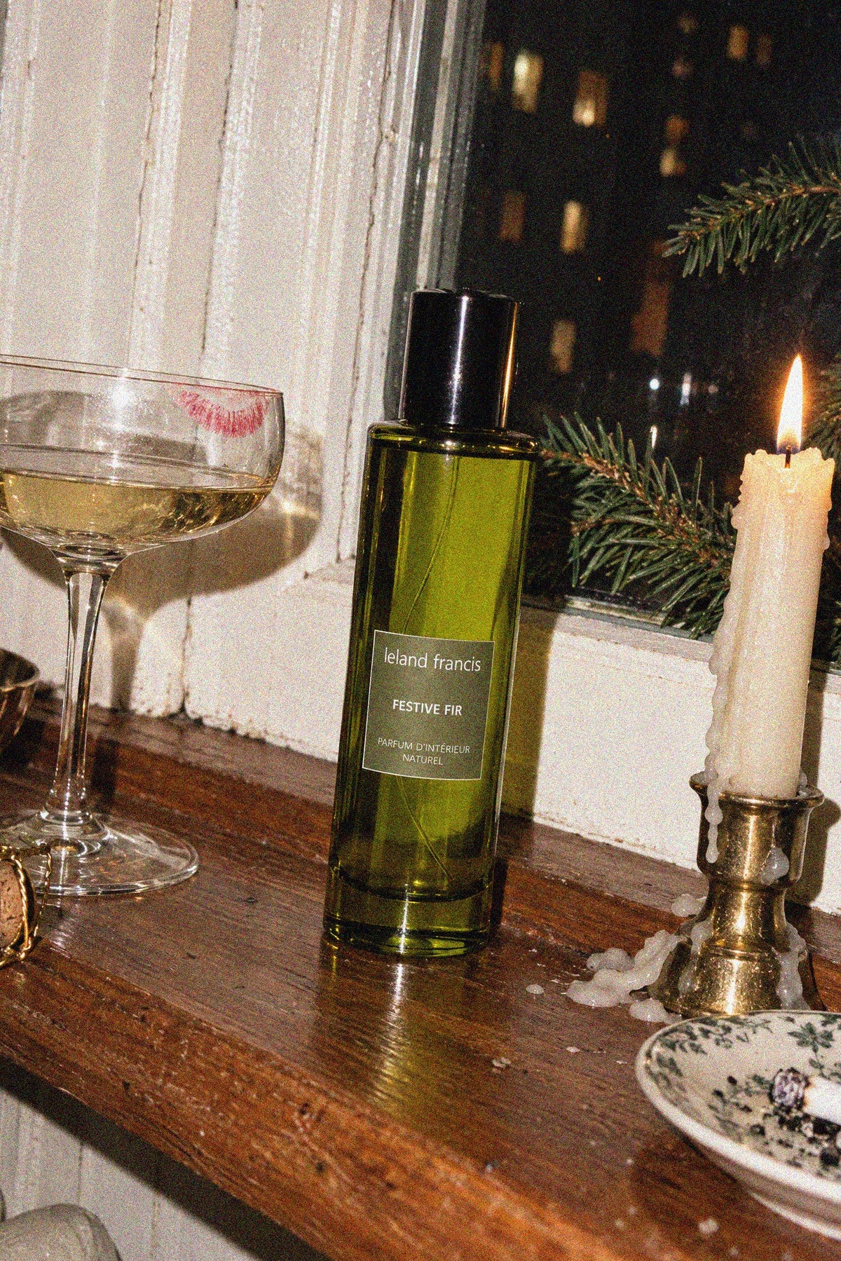 A green glass bottle of Leland Francis Festive Fir Room Spray stands on a wooden windowsill next to a lit candle, a filled coupe glass, and a smoking cigarette dish.