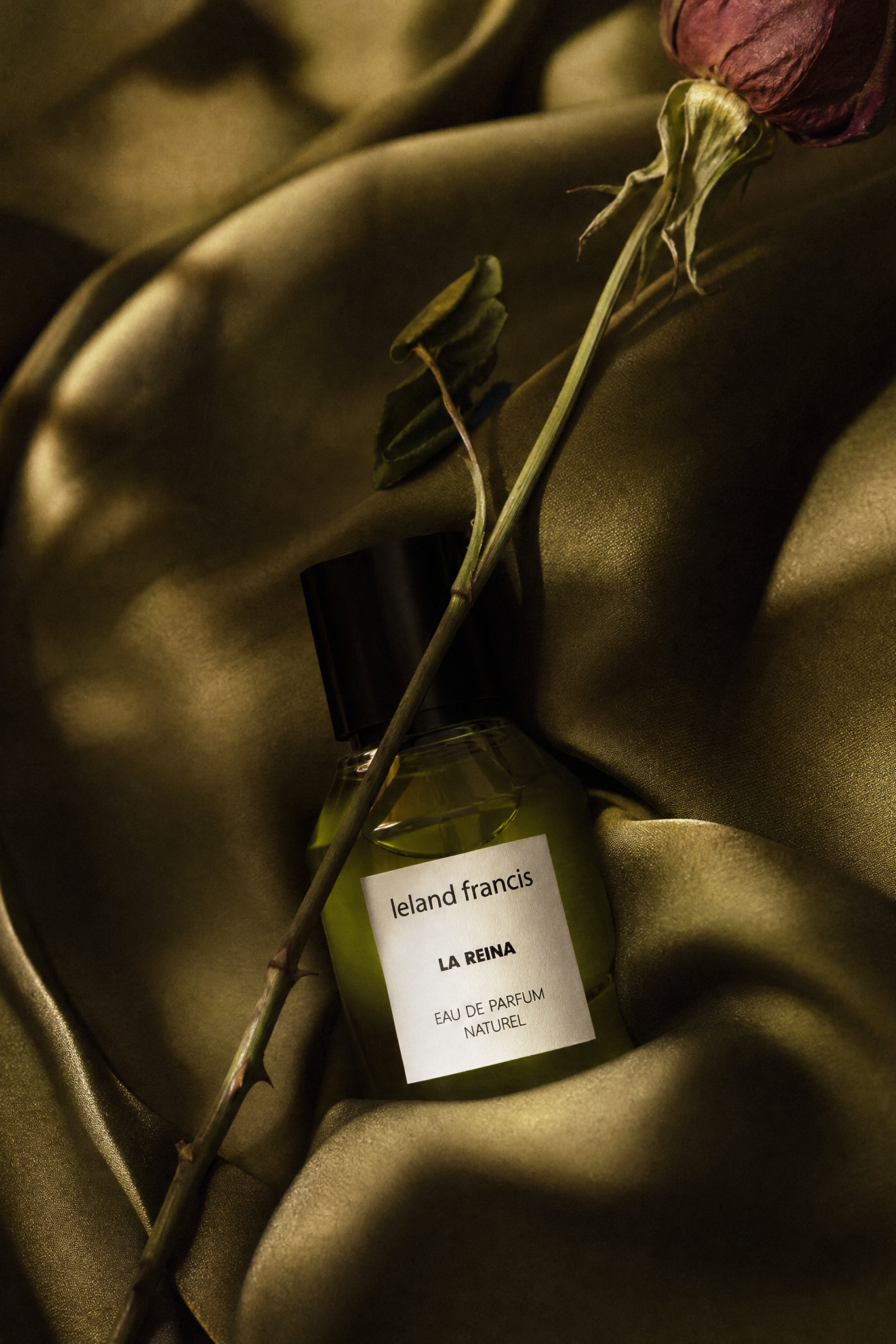 Perfume bottle labeled 'leland Francis La Reina' with a dried rose on a textured fabric background