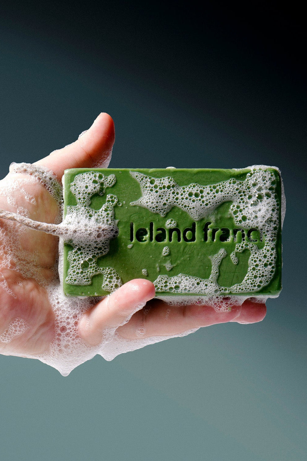 Green bar of soap with 'ileland frenn' branding held in a hand against a dark background