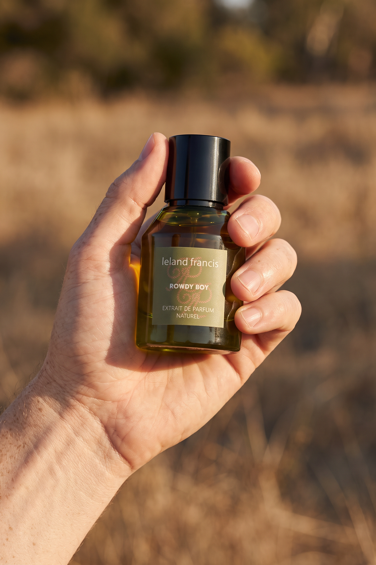 A hand holding a bottle of Leland Francis Rowdy Boy blurred outdoor dry grass background.