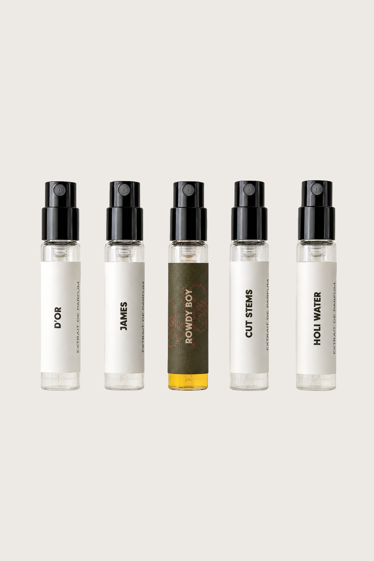 Five clear glass perfume spray bottles with black atomizers and white labels are lined up against a light beige background. The labels read D'OR, JAMES, ROWDY BOY , CUT STEMS, and HOLI WATER.