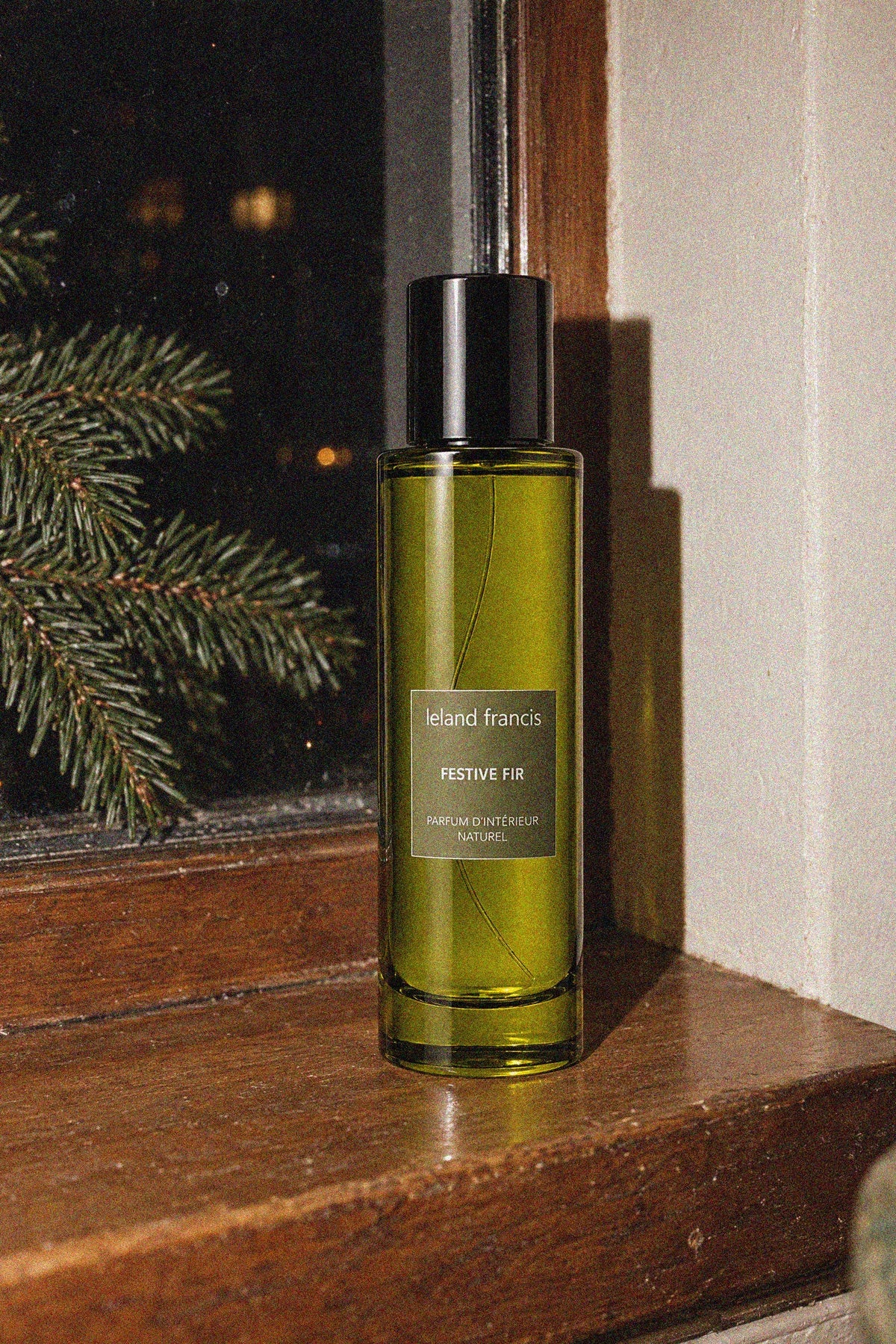 A bottle of Leland Francis Festive Fir room spray sitting on a wooden windowsill at night with a pine branch visible in the background.
