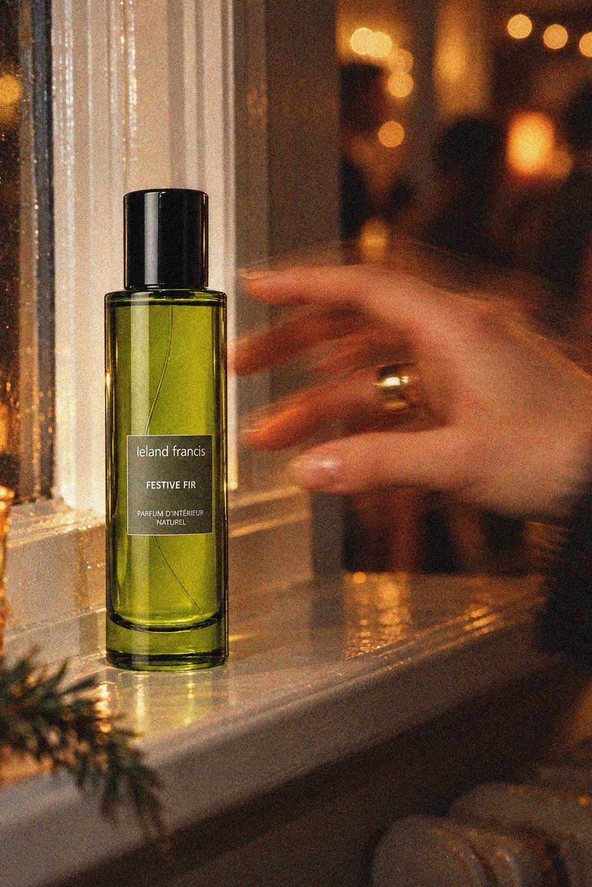 A hand reaches toward a green glass bottle of Leland Francis Festive Fir Room Spray  on a windowsill, with warm candlelight and blurred background lights.