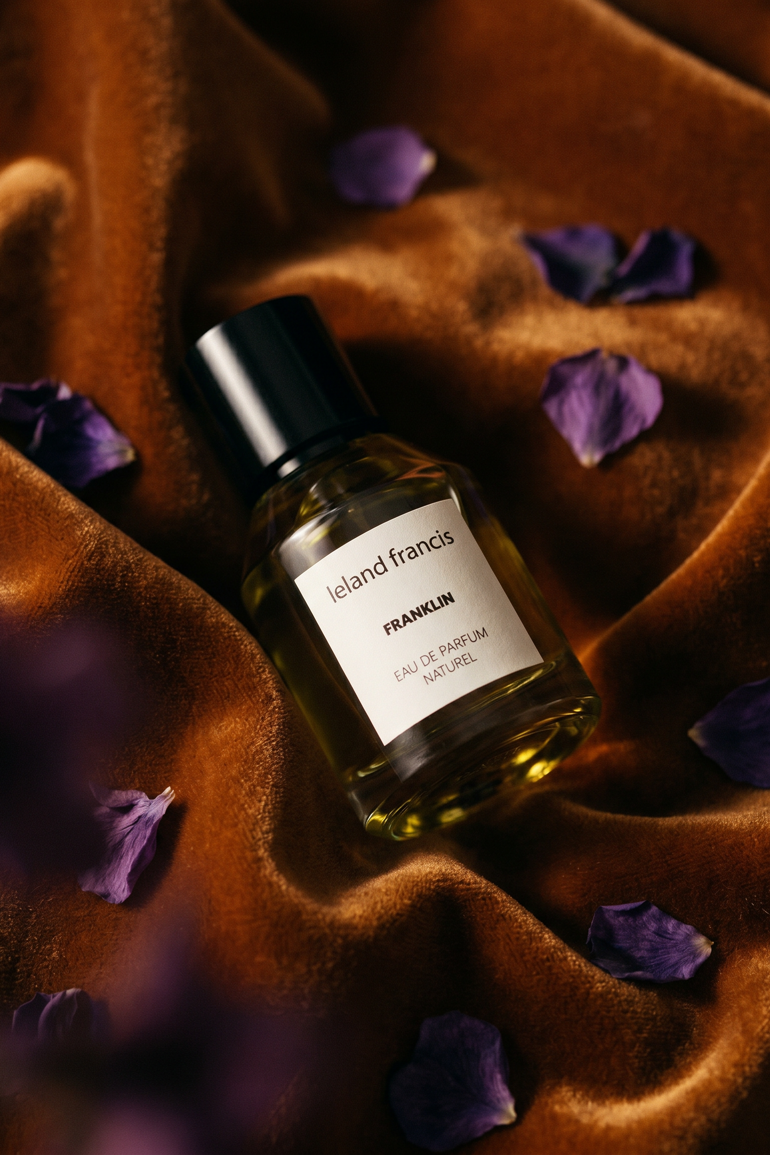 Perfume bottle labeled 'Leland Francis' on a textured brown surface with purple petals.