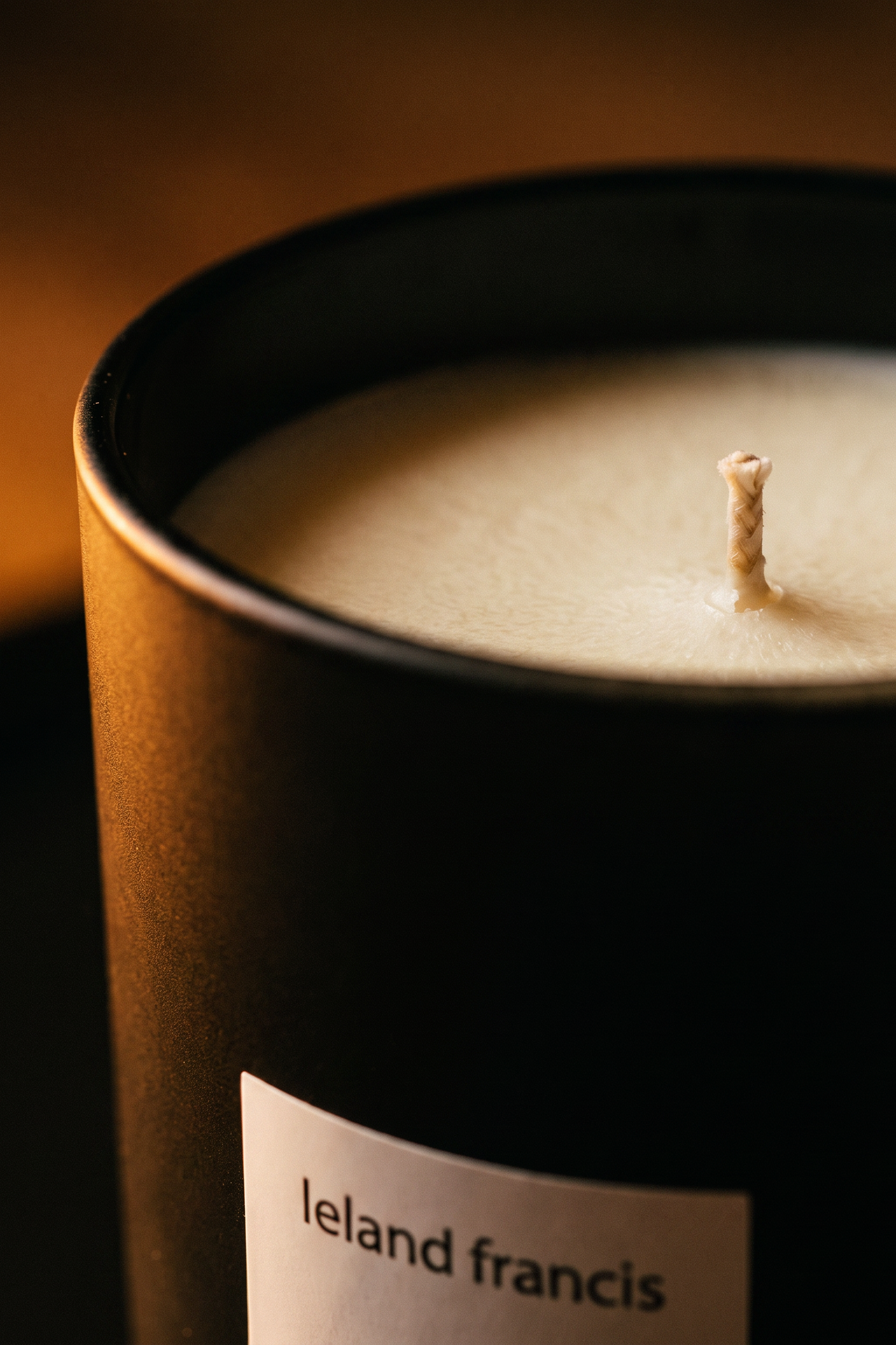 A close-up of a candle in a DARK GREEN container with a white label reading 'leland francis' and a single wick, lit with warm golden light against a dark background.