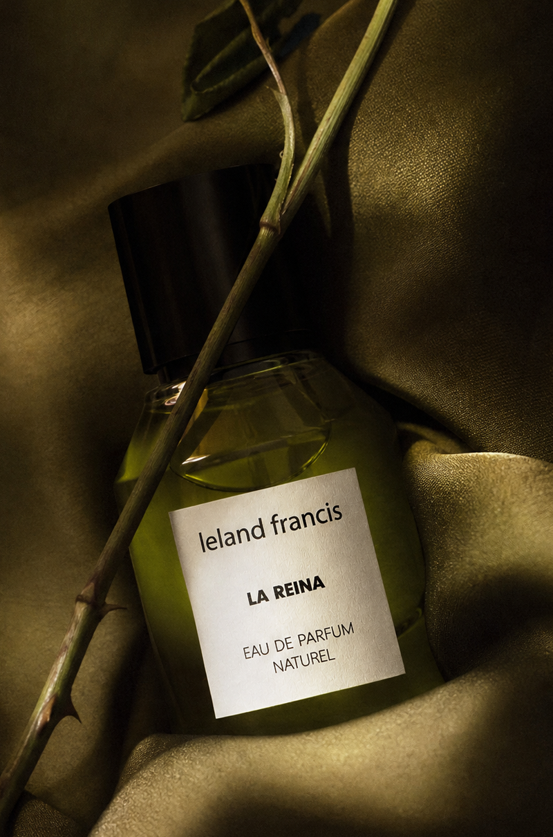 Perfume bottle labeled 'leland Francis La Reina' with a dried rose on a textured fabric background