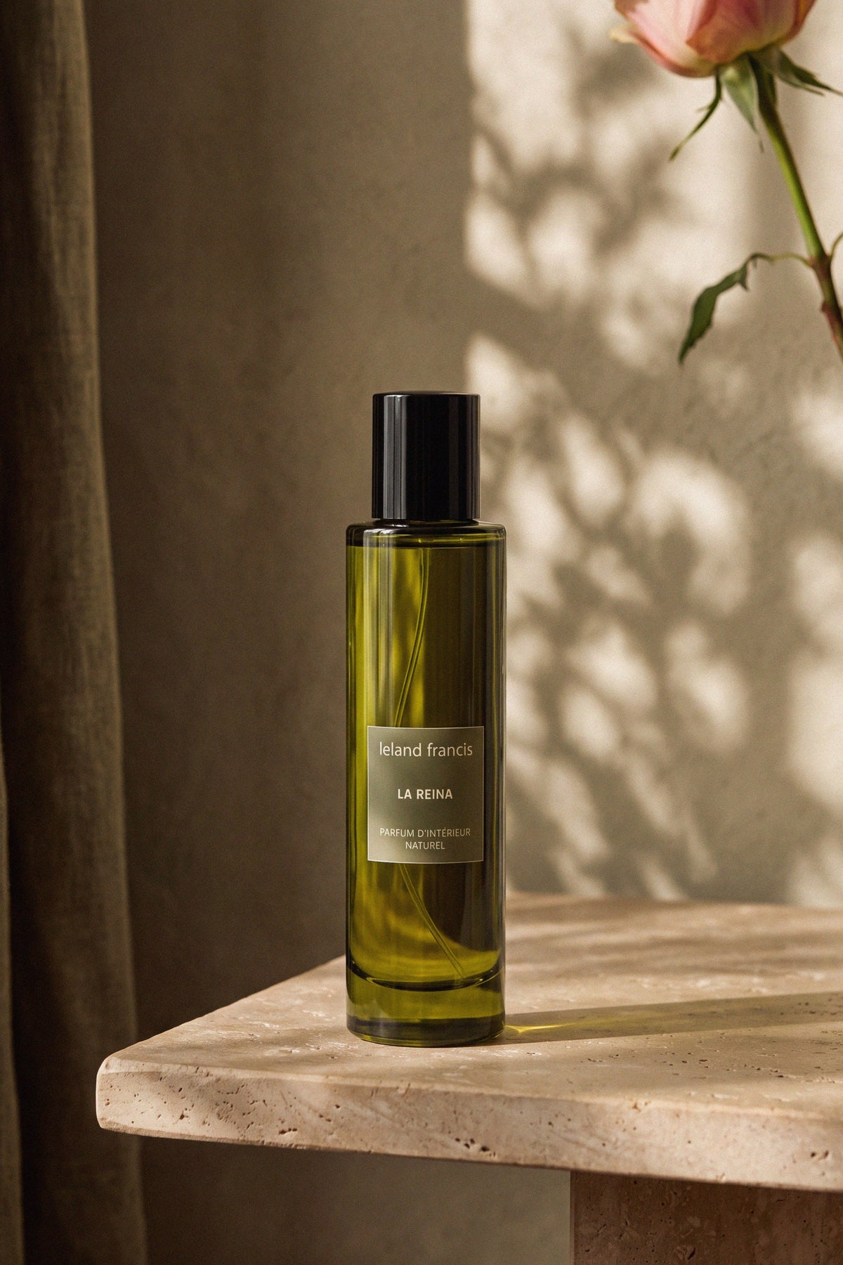 La Reina Room Spray on a travertine side table beside a single pink rose, afternoon light