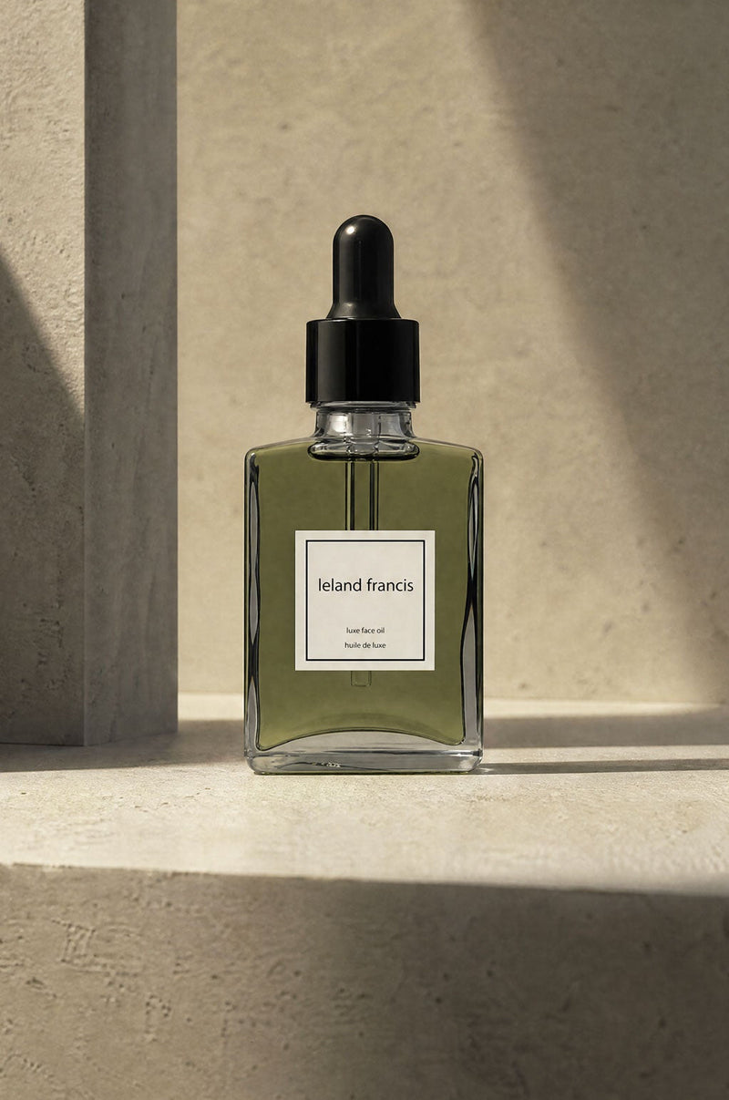 A rectangular glass dropper bottle with a black cap and a white label reading leland francis luxe face oil. stands on a stone surface in warm sunlight and shadow against a textured beige wall.
