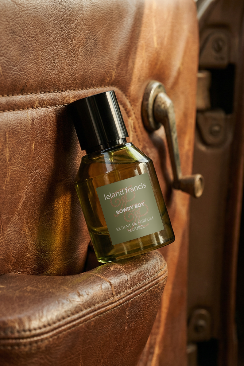 Green glass perfume bottle with a black cap and a muted green label reading 'Leland Francis Rowdy  Boy', resting in the armrest of a distressed brown leather chair.