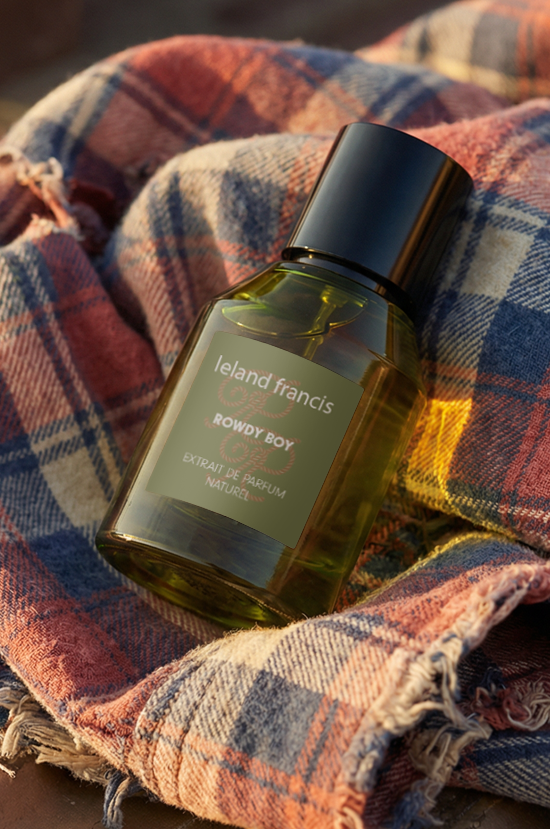 A bottle of Leland Francis Rowdy Boy rests on an old metal truck bed, wrapped in a fringed plaid shirt in warm autumnal tones.