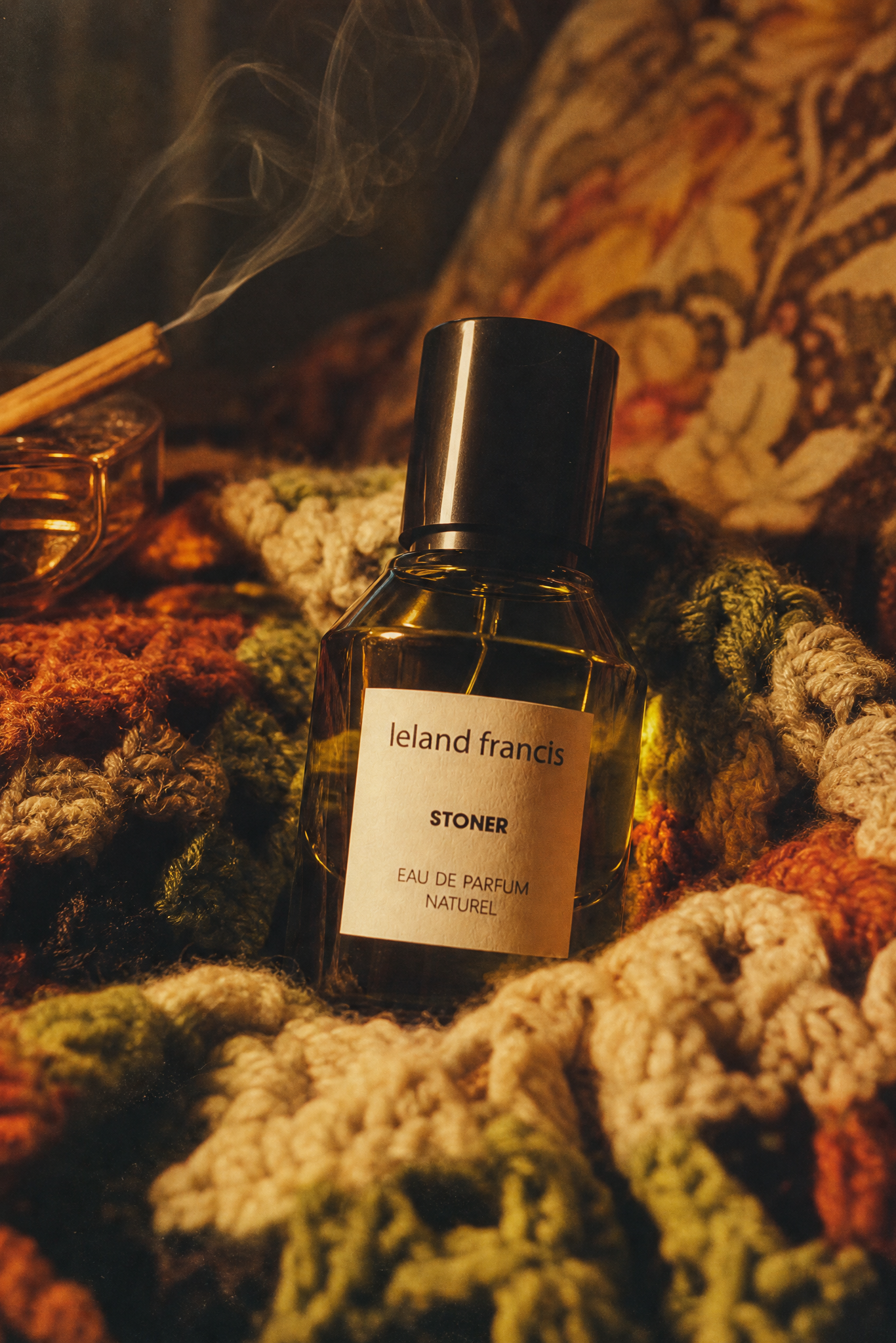 Perfume bottle labeled 'leland Francis' on a textured surface with smoke and patterned fabric in the background.